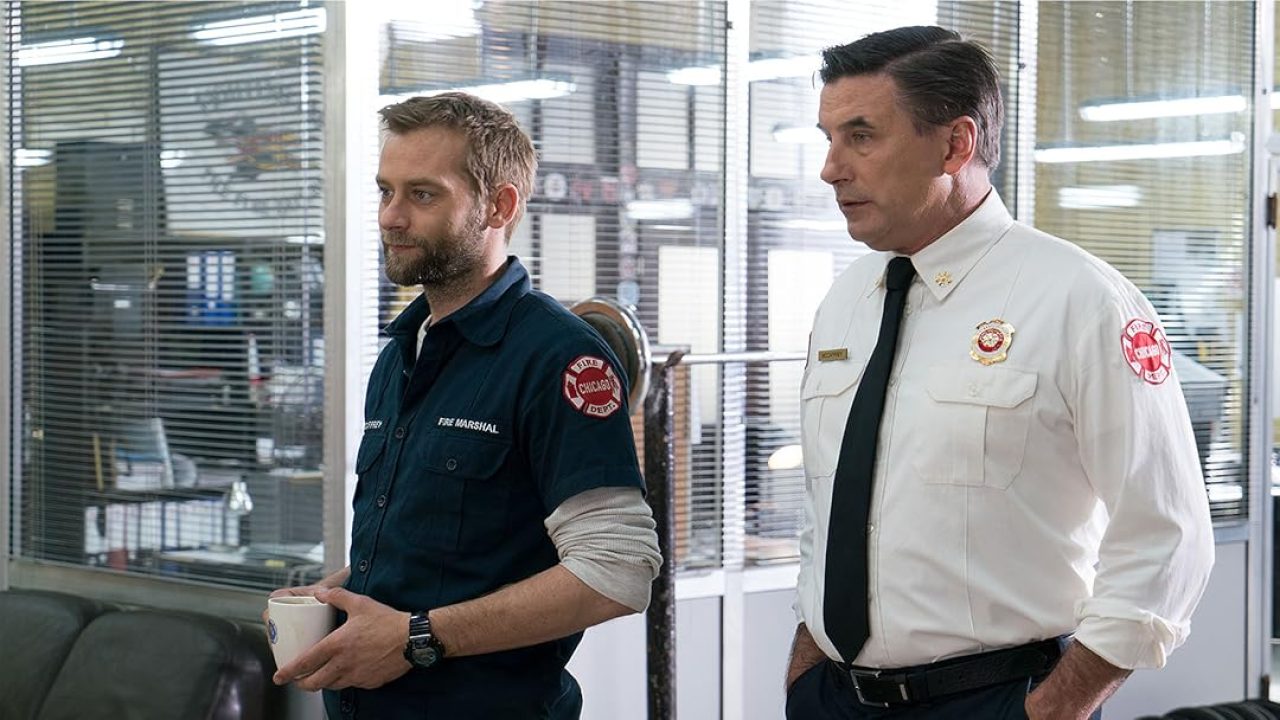 Dois homens adultos, um em uniforme azul de bombeiro com identificação de “fire marshal” e outro em camisa branca com gravata e distintivo, caminham lado a lado dentro de um escritório com paredes de vidro e persianas. Ambos têm expressão séria; o bombeiro segura uma caneca, e o ambiente sugere uma central administrativa ou quartel, com mesas e equipamentos ao fundo.