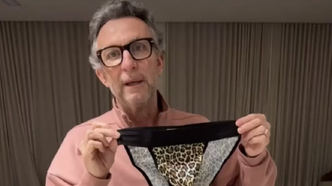 A imagem mostra um homem usando óculos e um suéter rosa, segurando uma peça de roupa íntima com estampa de onça. Ele está em um ambiente interno, com cortinas fechadas ao fundo, e parece estar falando ou explicando algo enquanto exibe o item diante da câmera