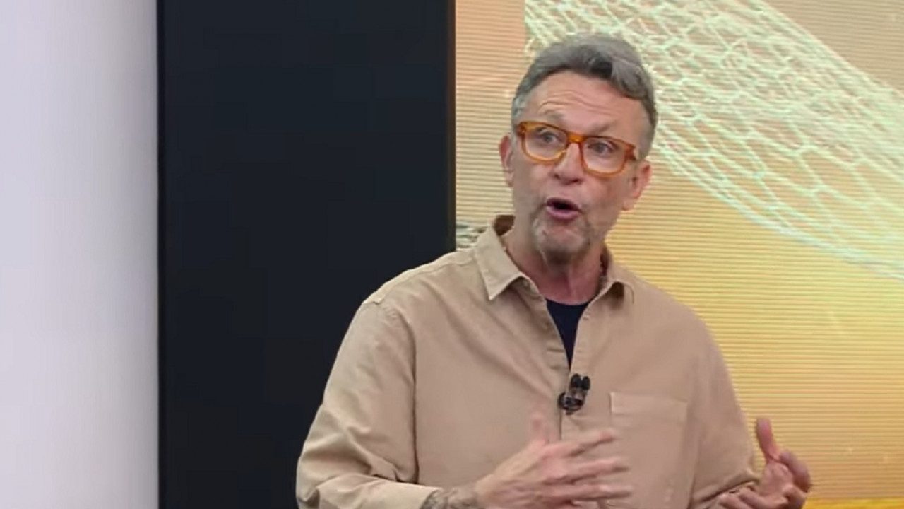 Homem de óculos com armação alaranjada aparece em estúdio de TV, gesticulando enquanto fala. Ele usa camisa bege e camiseta escura por baixo, com microfone preso ao peito. Ao fundo, há um painel iluminado com padrão gráfico abstrato em tons de amarelo