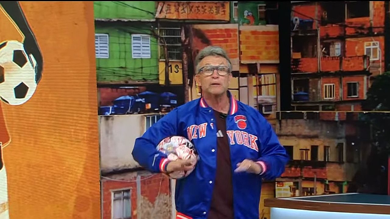 Homem usando óculos e uma jaqueta azul escrito “New York”, segurando uma bola de futebol em um estúdio de TV com cenário de casas ao fundo.