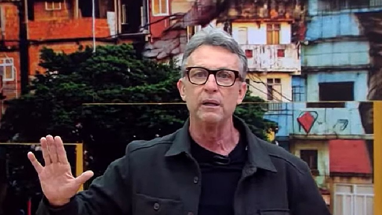 Apresentador levanta a mão durante fala em estúdio com imagem de comunidade ao fundo