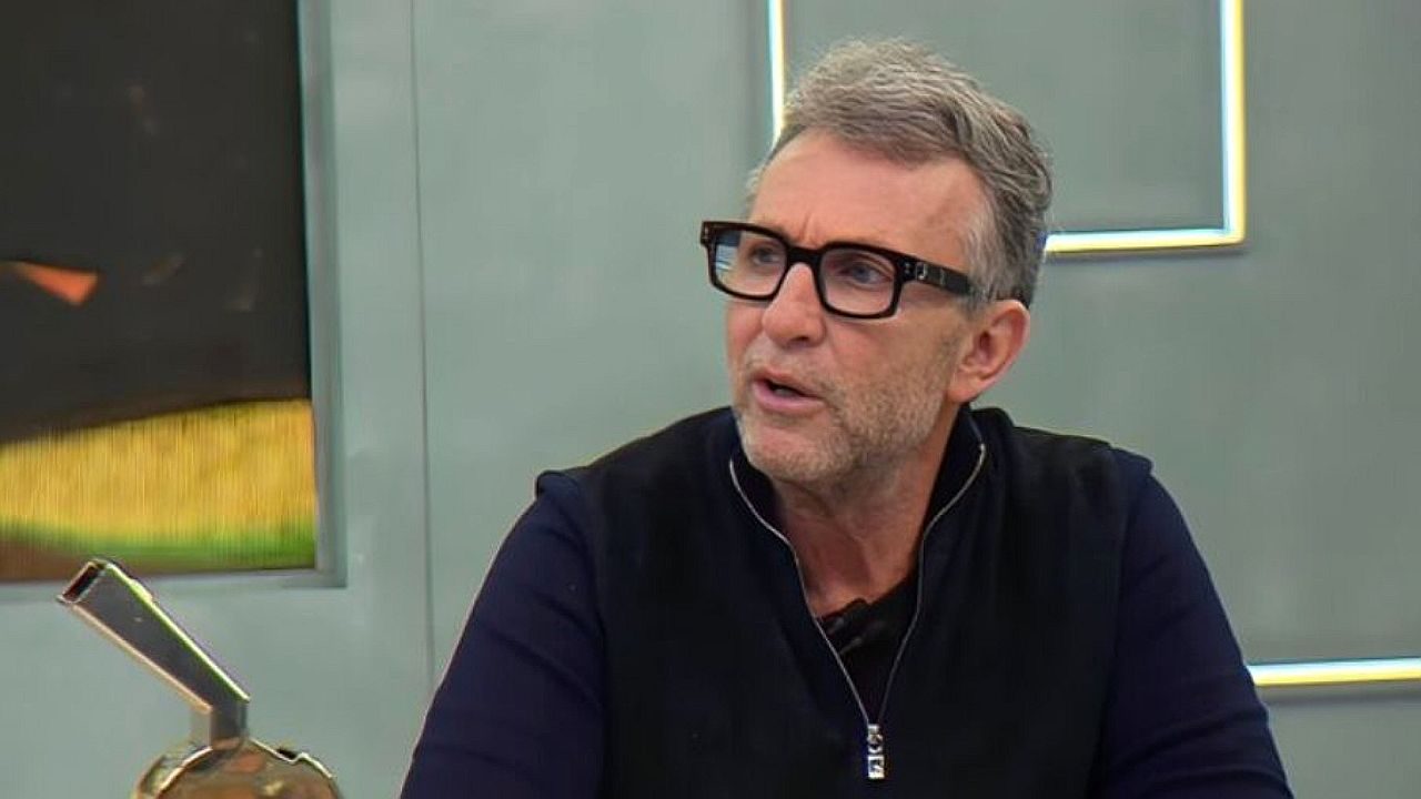 Um homem aparece em um estúdio, usando óculos de armação escura e roupa preta. Ele tem cabelo curto grisalho e barba por fazer. Está levemente virado para o lado, com expressão atenta, como se estivesse ouvindo ou respondendo a alguém durante uma conversa. Ao fundo, há elementos de cenário moderno com iluminação suave e linhas geométricas, típicos de programas de entrevista ou debate.