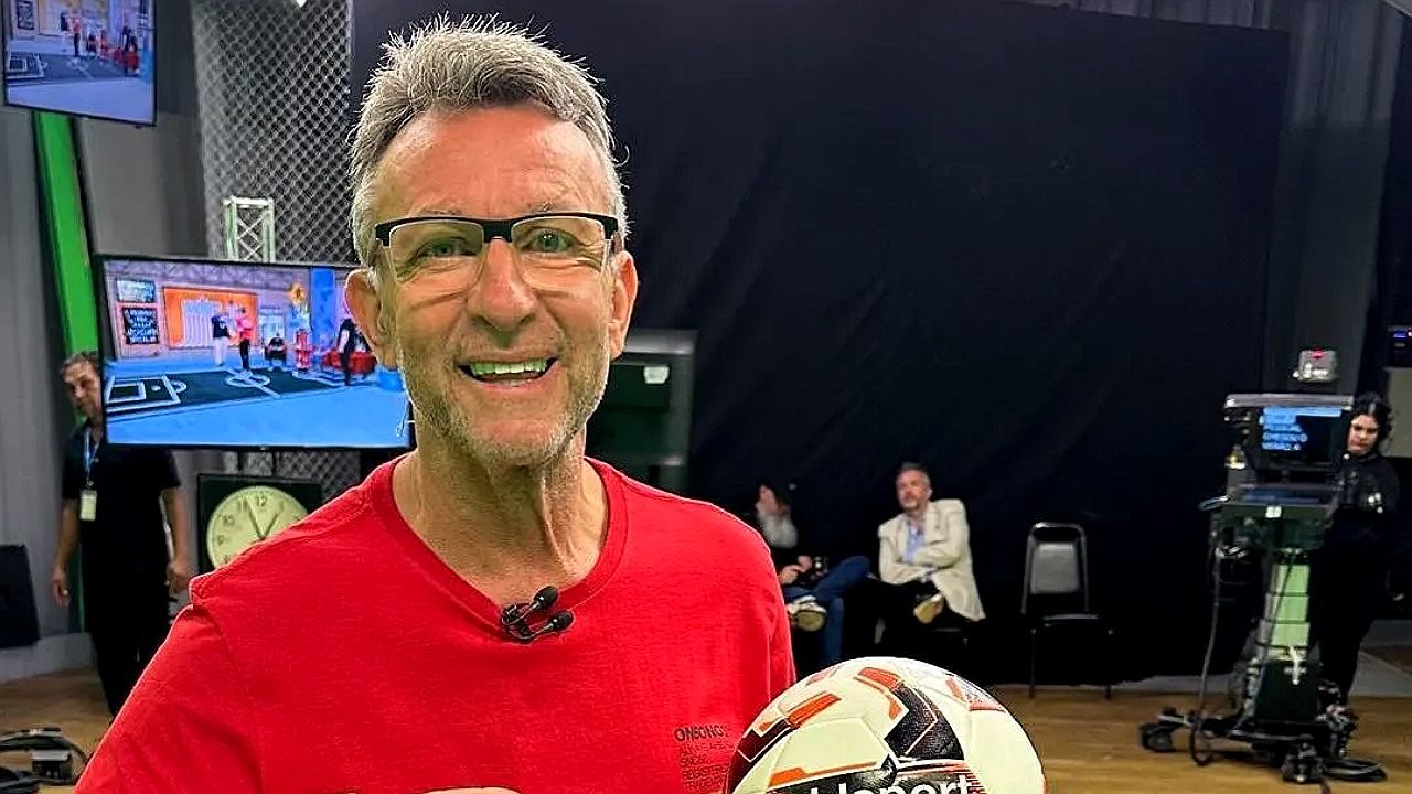 Apresentador esportivo segura bola de futebol em estúdio de TV com câmeras e equipe ao fundo