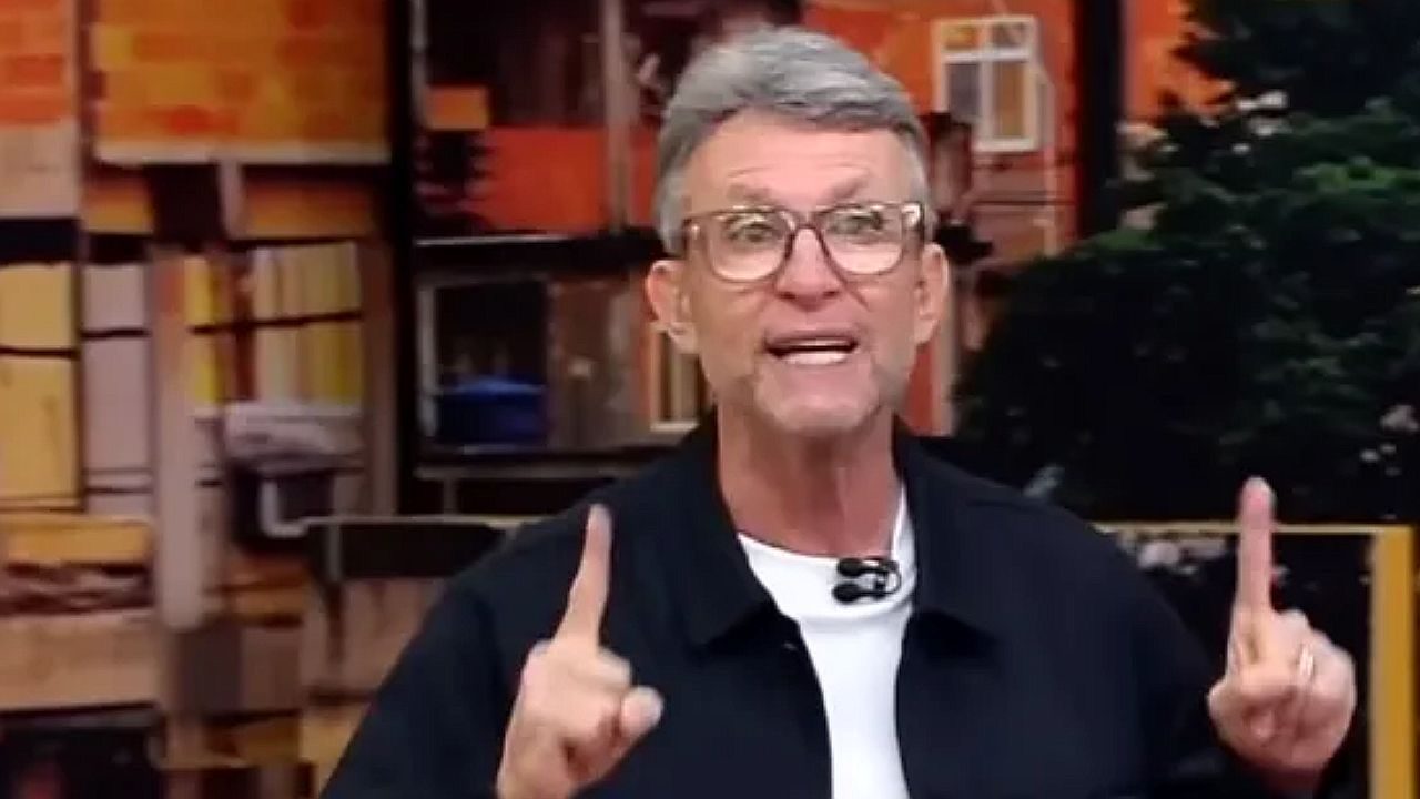Homem de óculos aponta para cima durante apresentação em programa de TV no estúdio