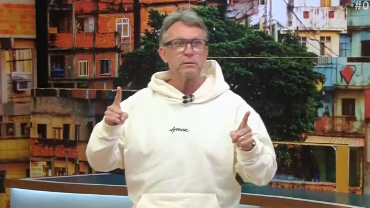 Craque Neto de moletom branco faz gesto com os dedos apontados em estúdio com painel de comunidade ao fundo