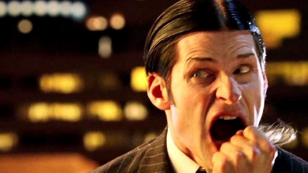 Homem de terno escuro aparece em close com expressão exagerada, boca aberta como se estivesse gritando ou reagindo de forma dramática. O fundo desfocado com luzes urbanas noturnas sugere cena estilizada de filme ou série ambientada à noite.