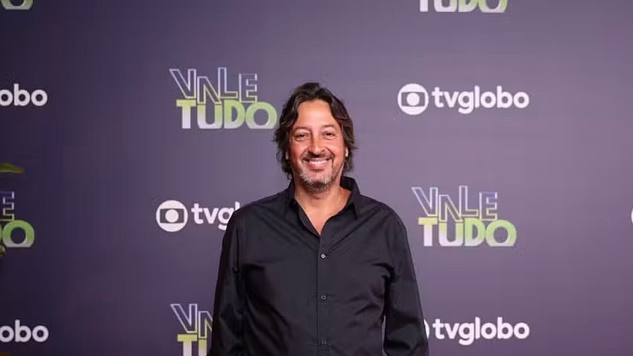 Ator posa sorrindo em evento da novela Vale Tudo com logos da TV Globo ao fundo