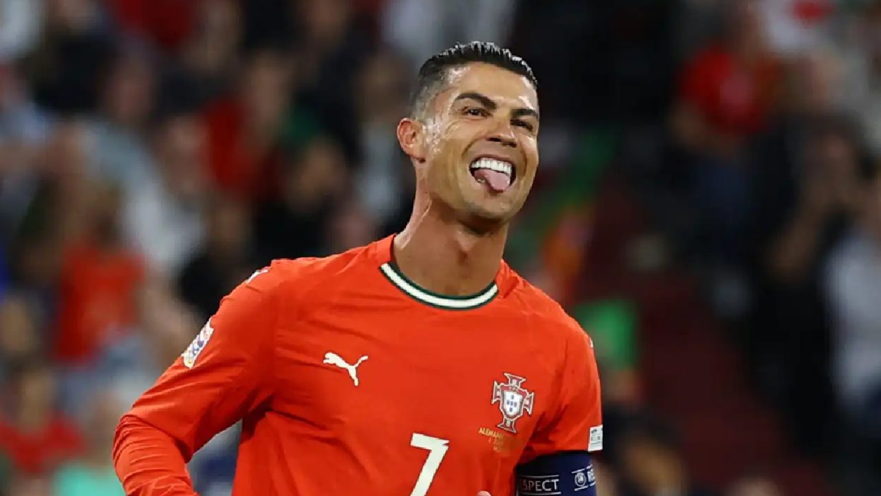 Jogador de futebol com uniforme vermelho da seleção de Portugal comemora em campo, sorrindo e mostrando a língua, com torcida desfocada ao fundo