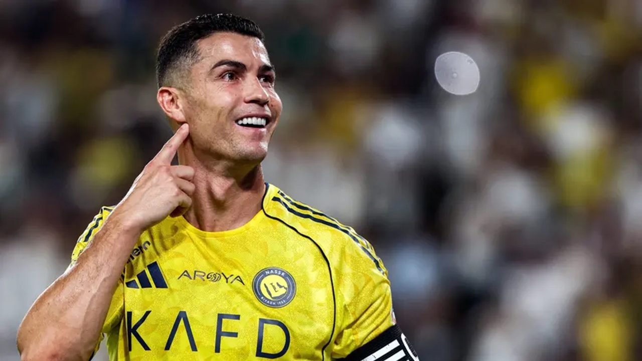 Cristiano Ronaldo com uniforme amarelo sorri e aponta para o rosto em campo, com arquibancada desfocada ao fundo, durante partida noturna