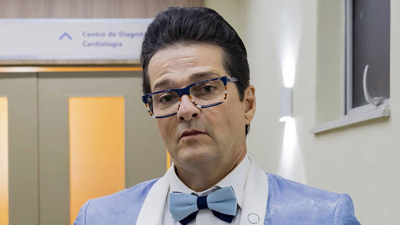 Marcelo Serrado como Crô na novela Três Graças usa paletó azul claro com gravata borboleta e óculos coloridos no corredor de hospital