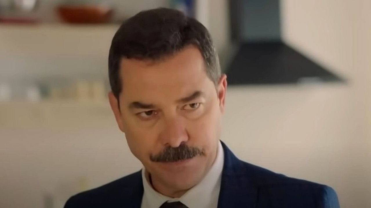 Homem de terno escuro e gravata aparece em ambiente interno, com expressão séria e olhar atento. Ele tem bigode bem aparado e está diante de cenário doméstico ao fundo, sugerindo cena de filme ou série dramática.