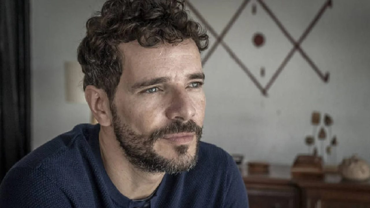 Homem de barba e cabelo curto e cacheado olha pensativo para o lado em ambiente interno, com parede decorada e iluminação suave ao fundo