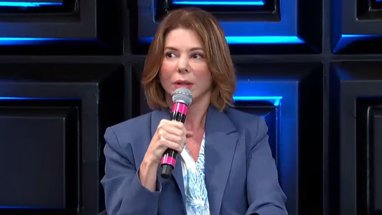 Daniela Abravanel segura microfone e fala durante participação em coletiva com cenário iluminado em azul ao fundo