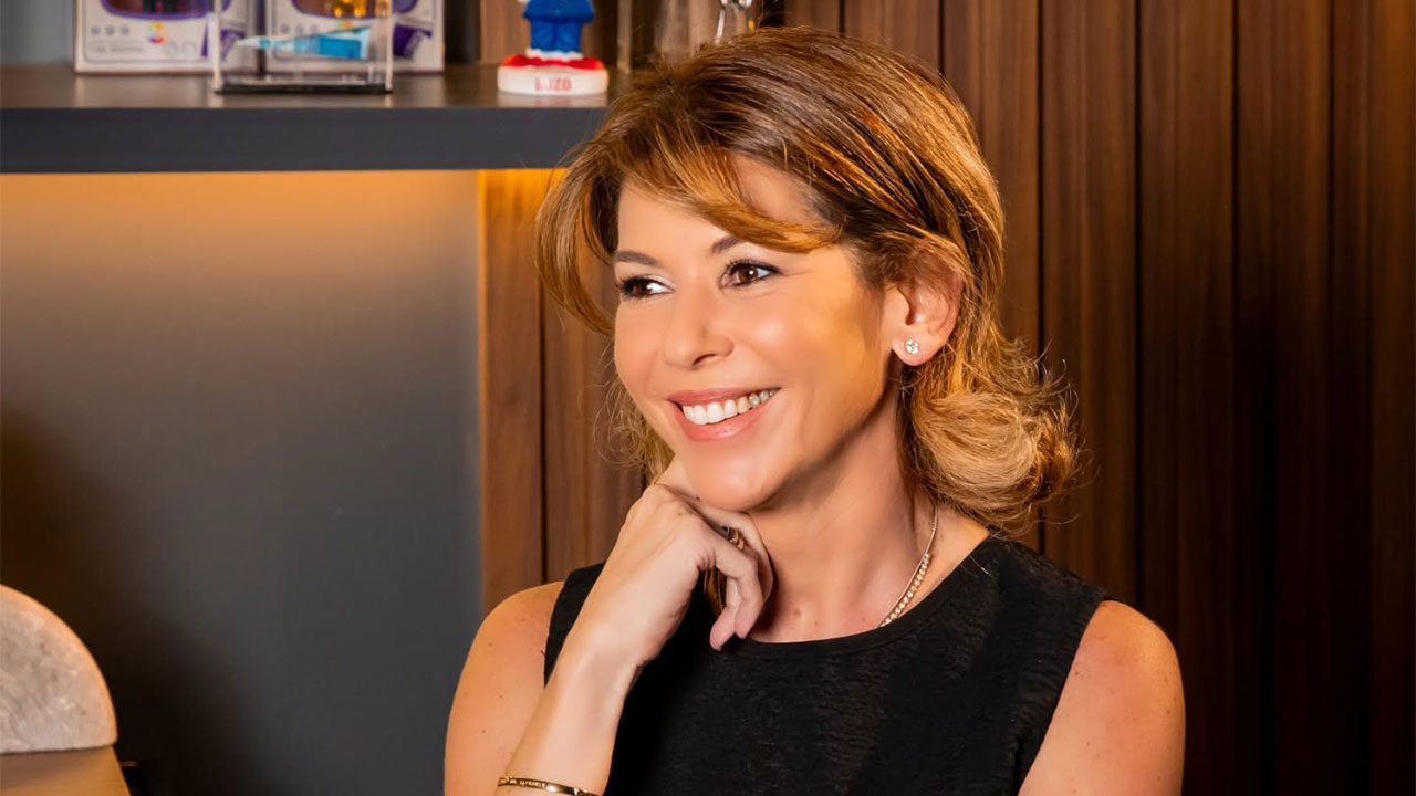 Daniela Beyruti com cabelo castanho solto, sorrindo e com a mão direita no queixo, posando em escritório com painel de madeira ao fundo