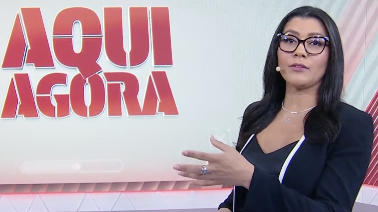 Daniele Brandi, de óculos e blazer escuro, gesticula em estúdio diante do painel digital com a marca do Aqui Agora