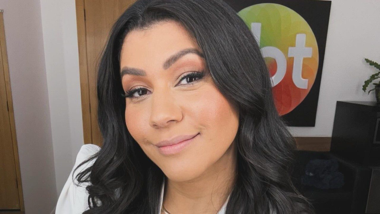 Daniele Brandi sorri para a câmera com blazer branco diante de painel do SBT ao fundo