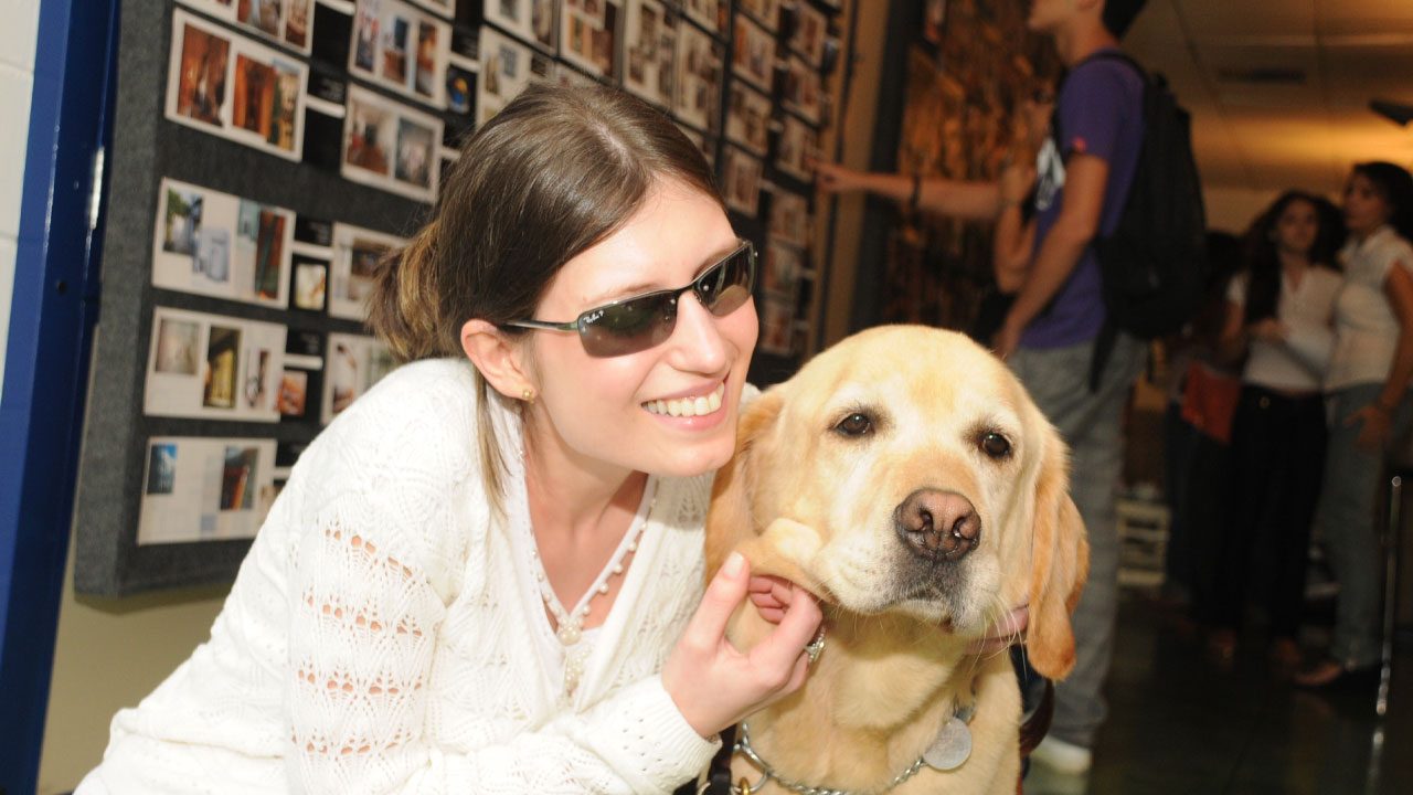 Danieli Haloten sorrindo ao lado de seu cão-guia labrador em ambiente interno com murais ao fundo