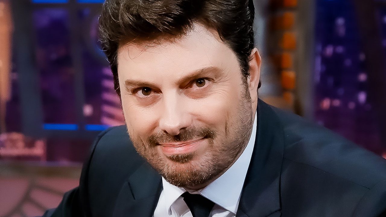 O apresentador Danilo Gentili sorri com terno preto e gravata, olhando para a câmera no estúdio do talk show The Noite, no SBT