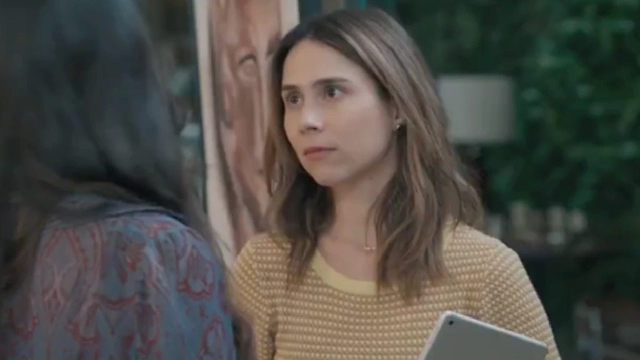 A imagem mostra uma mulher de cabelos castanhos e expressão séria, segurando um tablet. Ela veste uma blusa amarela clara com textura e está de frente para outra pessoa, que aparece de costas