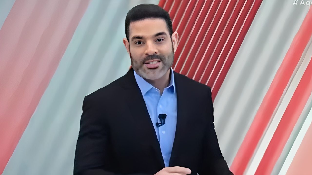 A imagem mostra Darlisson Dutra, um homem de terno escuro e camisa azul clara, usando microfone de lapela. Ele está diante de um fundo com faixas diagonais em tons de vermelho e branco, que serve de cenário para o programa de televisão Aqui Agora