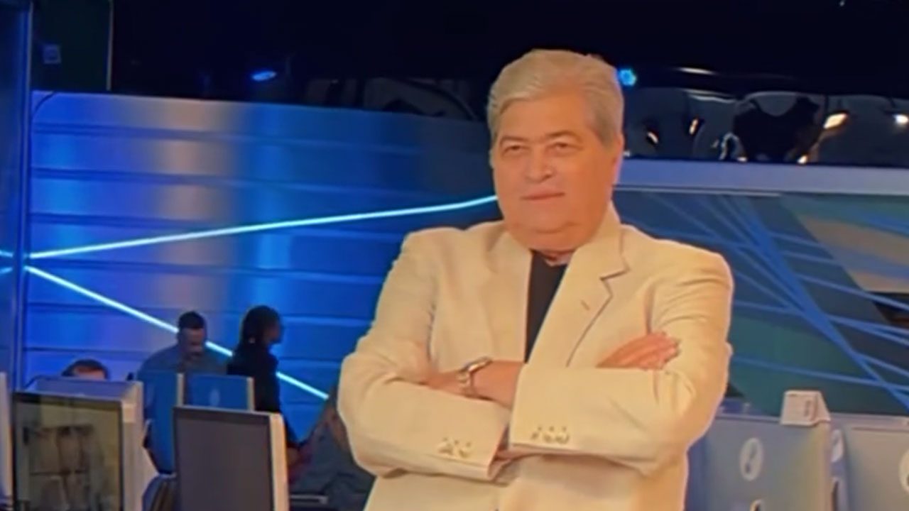 Datena de braços cruzados em frente aos computadores na redação da RedeTV! com cenário azul iluminado ao fundo