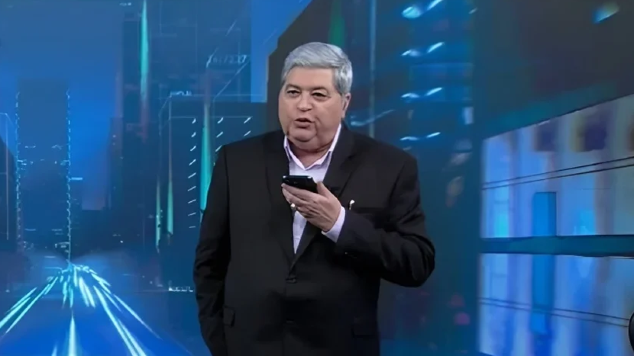 José Luiz Datena apresentando o Tá na Hora com celular na mão e cenário virtual ao fundo
