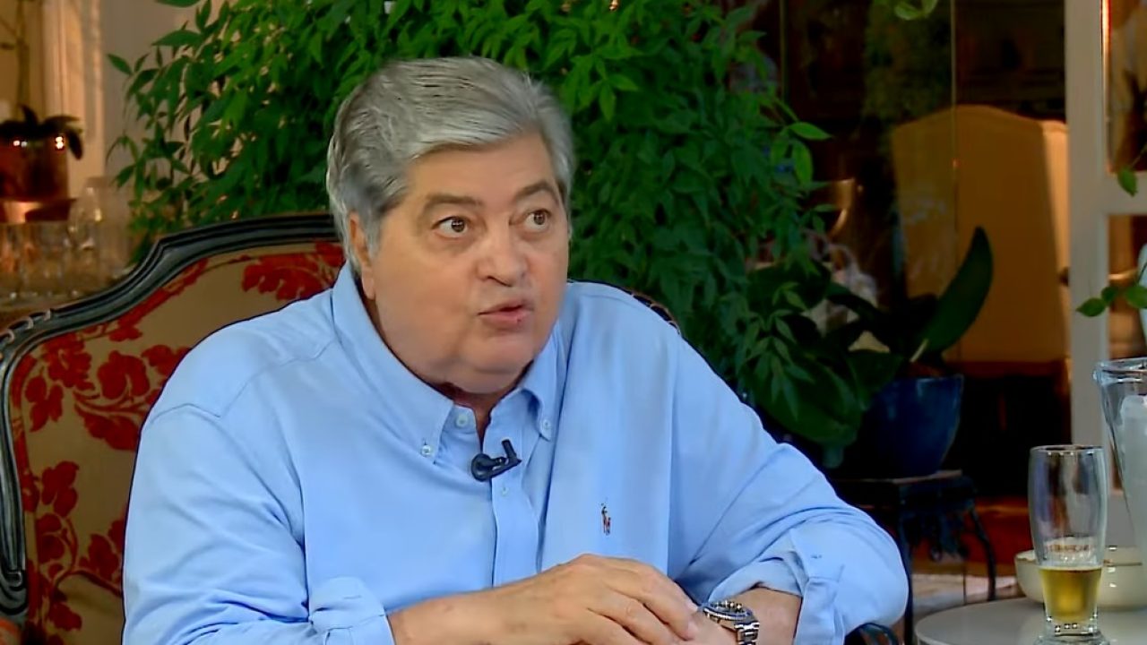 José Luiz Datena sentado em entrevista com camisa azul e copo de bebida sobre a mesa