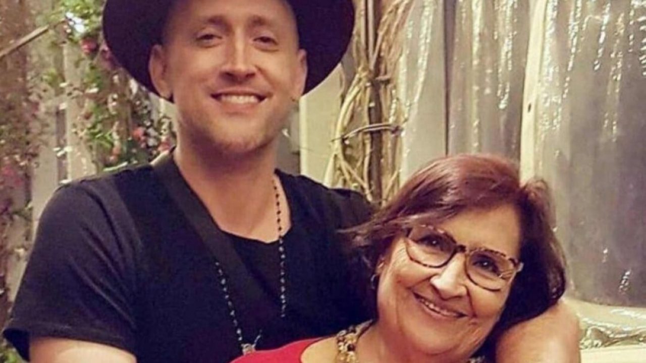 Na imagem, o ator Paulo Gustavo aparece abraçado com a mãe, Déa Lúcia, em um momento de afeto e descontração. Sorridente, Paulo veste camiseta preta, chapéu escuro e um colar com contas, transmitindo seu estilo característico. Déa também sorri, usando óculos de grau e colar dourado, com os cabelos curtos e escuros.