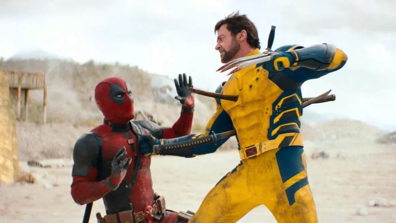 Cena de ação mostra Deadpool, em seu traje vermelho e preto, sendo empurrado para trás por Wolverine, que veste uniforme amarelo e azul. Wolverine ataca com garras de adamantium estendidas enquanto ambos lutam em um deserto claro, com estruturas de madeira e montanhas ao fundo