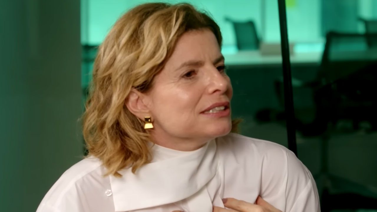 Debora Bloch sentada e falando durante entrevista em ambiente corporativo com fundo esverdeado