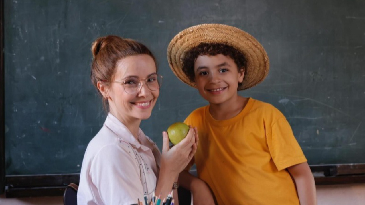 Débora Falabella sorri ao lado de menino com chapéu de palha em sala de aula segurando uma maçã em cena de novela