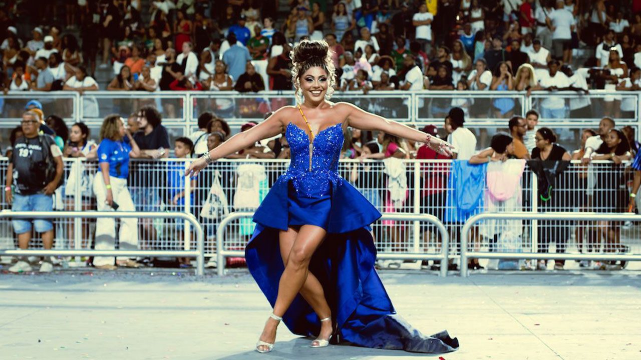 Deborah Caetano desfila no sambódromo com vestido azul rodado e braços abertos diante de arquibancadas lotadas durante o Carnaval