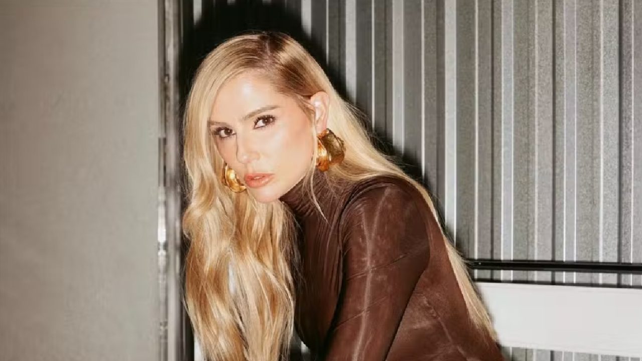 A mulher está posando com expressão séria, usando um body marrom justo e maxi-brincos dourados. Ela tem cabelos longos e loiros, penteados para a frente, e está apoiada levemente para frente em um ambiente interno com paredes metálicas ao fundo, em clima de ensaio fotográfico