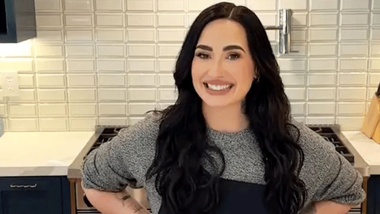 Uma mulher está em uma cozinha moderna, sorrindo diretamente para a câmera. Ela tem cabelos longos e escuros, usa uma blusa de manga longa cinza e um avental preto. Está com as mãos apoiadas na cintura, em uma postura confiante e descontraída. Ao fundo, há um fogão, bancada clara e uma parede com azulejos brancos em padrão geométrico, sugerindo um ambiente organizado e bem iluminado, típico de gravações culinárias.