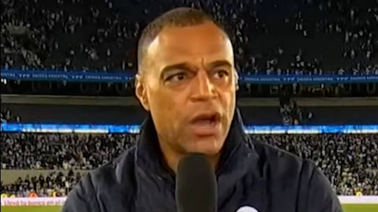 Comentarista esportivo fala ao microfone em estádio lotado durante transmissão de partida de futebol