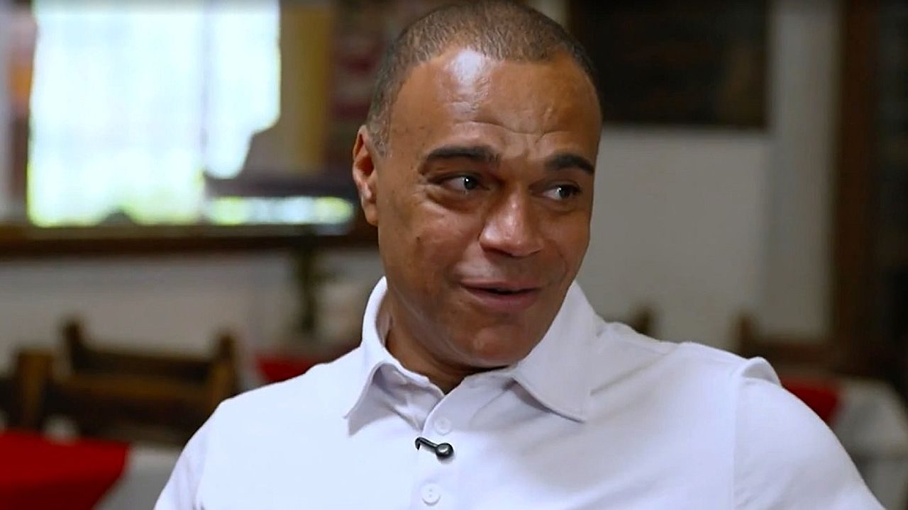 Denílson durante entrevista em restaurante usando camisa branca e microfone preso à gola