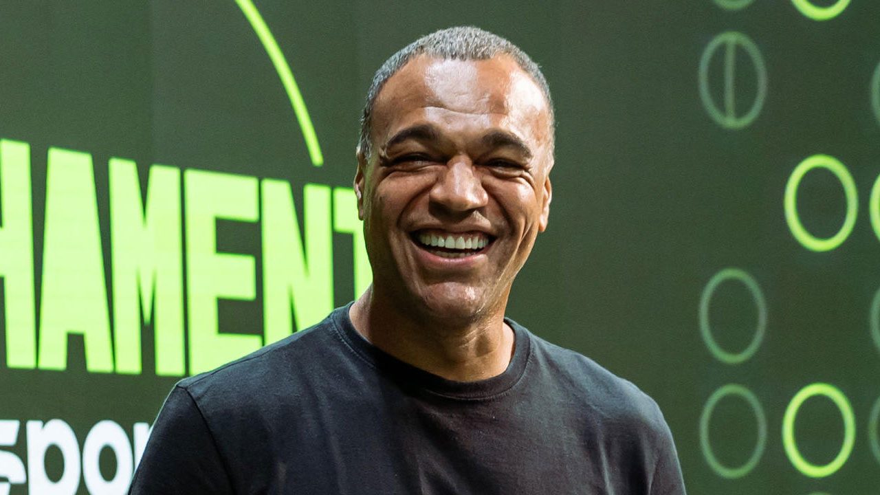 Denílson sorri vestindo uma camiseta preta básica em frente ao telão do Fechamento SporTV com o logo do programa