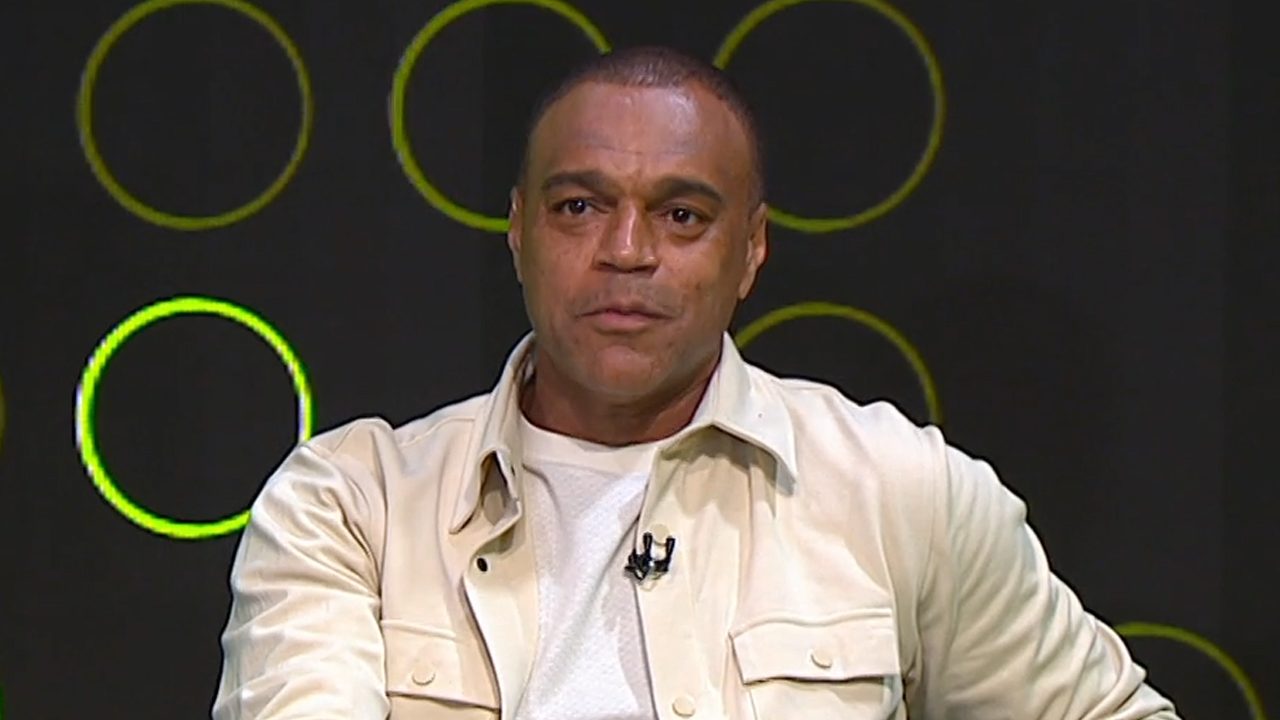 Denílson com jaqueta clara e camiseta branca falando durante participação em programa de TV com fundo escuro e círculos verdes