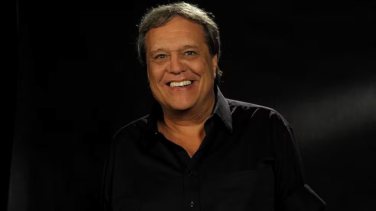 Dennis Carvalho sorri em retrato com fundo escuro durante gravação em estúdio de televisão