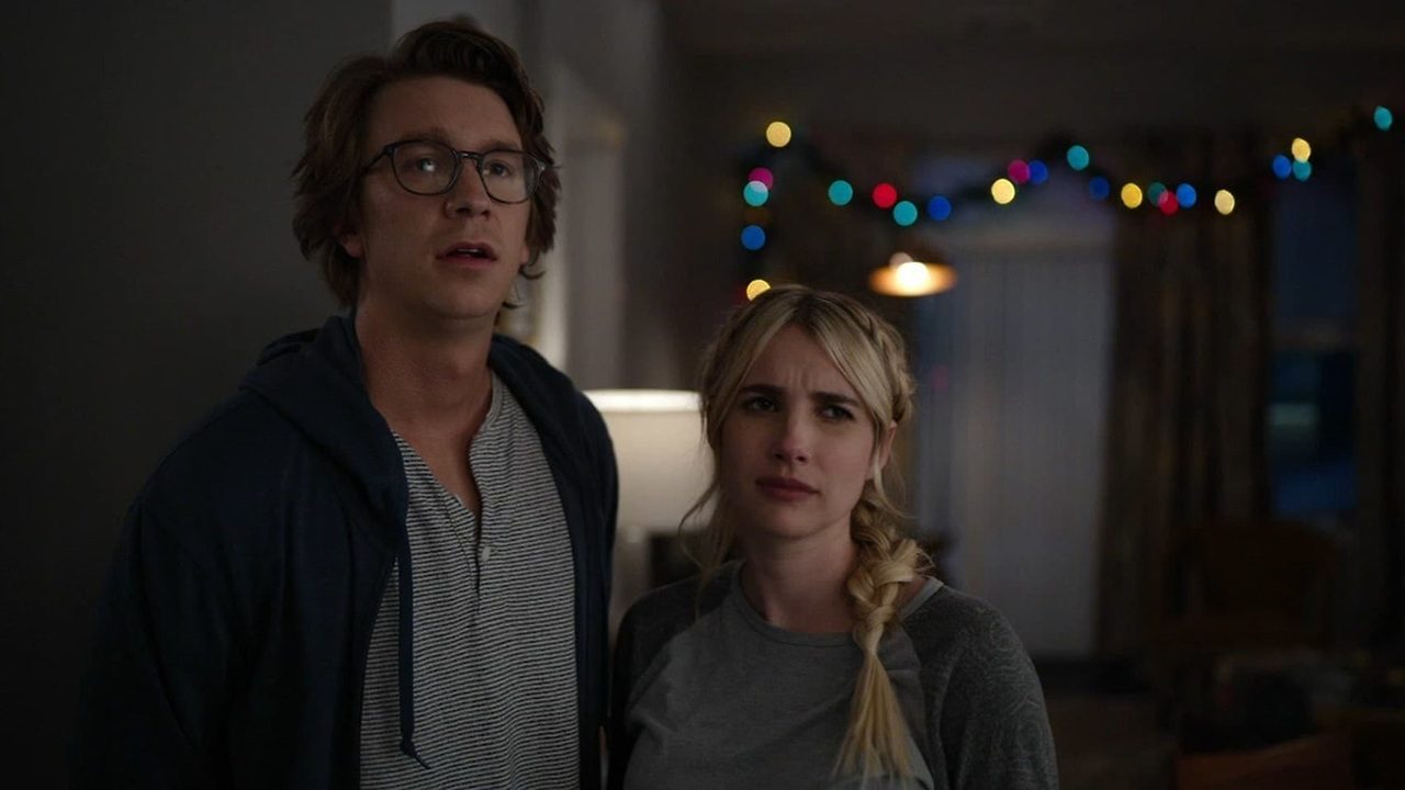 Homem de óculos e mulher de trança loira olham para frente com expressões de surpresa e confusão em ambiente interno decorado com luzes coloridas ao fundo