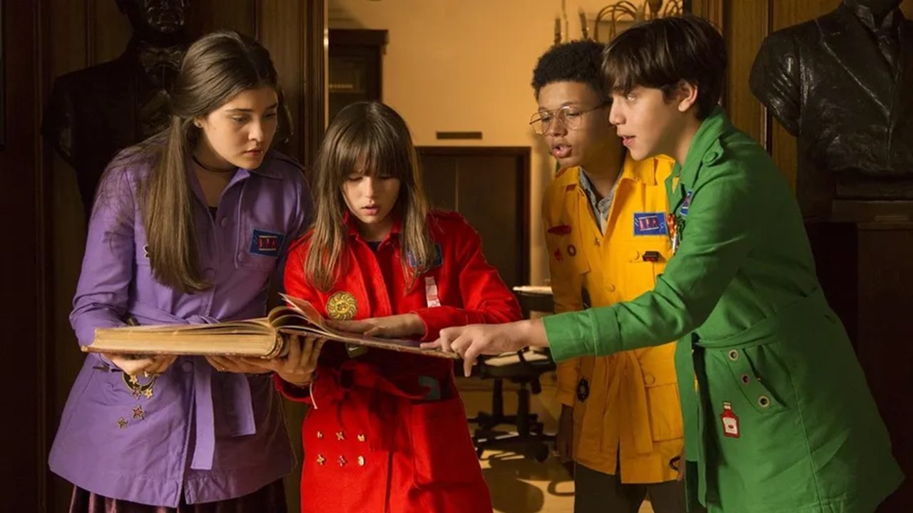 Quatro adolescentes, cada um usando um casaco de cor vibrante (roxo, vermelho, amarelo e verde), se reúnem em uma sala antiga e elegante enquanto observam com atenção um grande livro aberto. Eles parecem concentrados e curiosos, como se estivessem investigando algo importante. Ao fundo, há bustos de esculturas e móveis de madeira escura, reforçando um clima de mistério