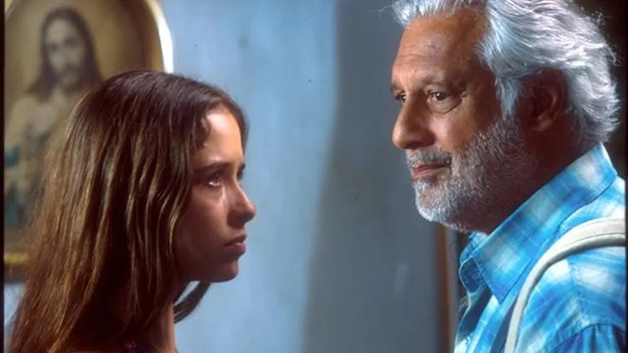 Homem de cabelos e barba brancos, vestindo camisa xadrez azul, encara uma mulher de cabelos longos castanhos que o observa com expressão séria. Ao fundo, há uma parede azul clara e um quadro religioso com a imagem de Jesus Cristo.