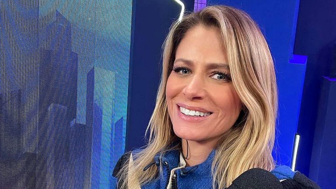 Apresentadora loira sorri diante de câmera em estúdio com iluminação azul ao fundo