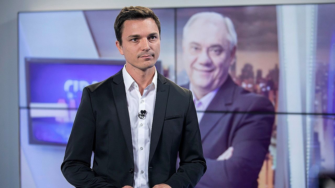 Jornalista Diego Rezende apresenta telejornal em estúdio com imagem de comentarista exibida ao fundo