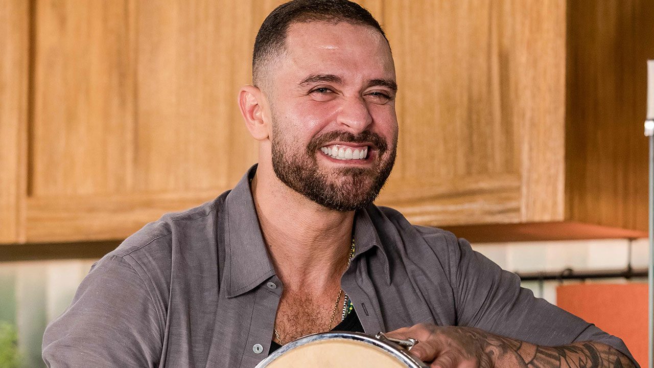Diogo Nogueira sorri com barba aparada e camisa cinza enquanto segura um instrumento musical em cenário de madeira clara