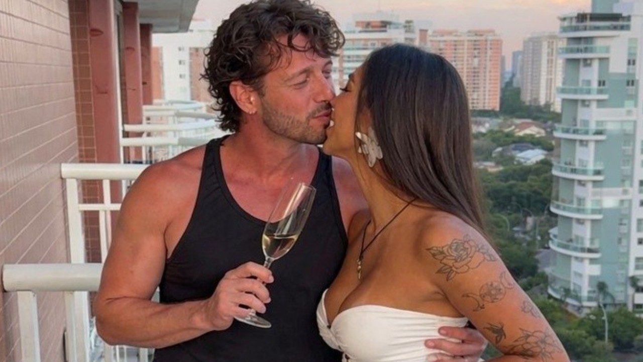 Diogo Venturieri e Didy Reis se beijam em varanda de apartamento enquanto seguram taça de espumante