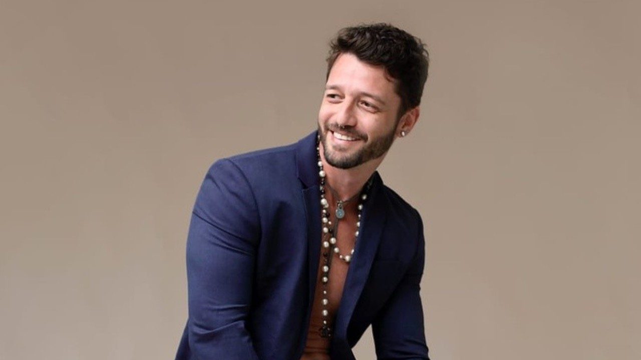 Diogo Venturieri sorri usando blazer azul aberto e colares posando em ensaio com fundo neutro
