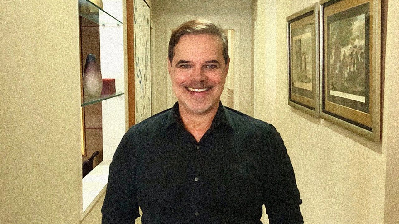 Diogo Vilela sorrindo em corredor decorado com quadros na parede e prateleira com vaso de cerâmica