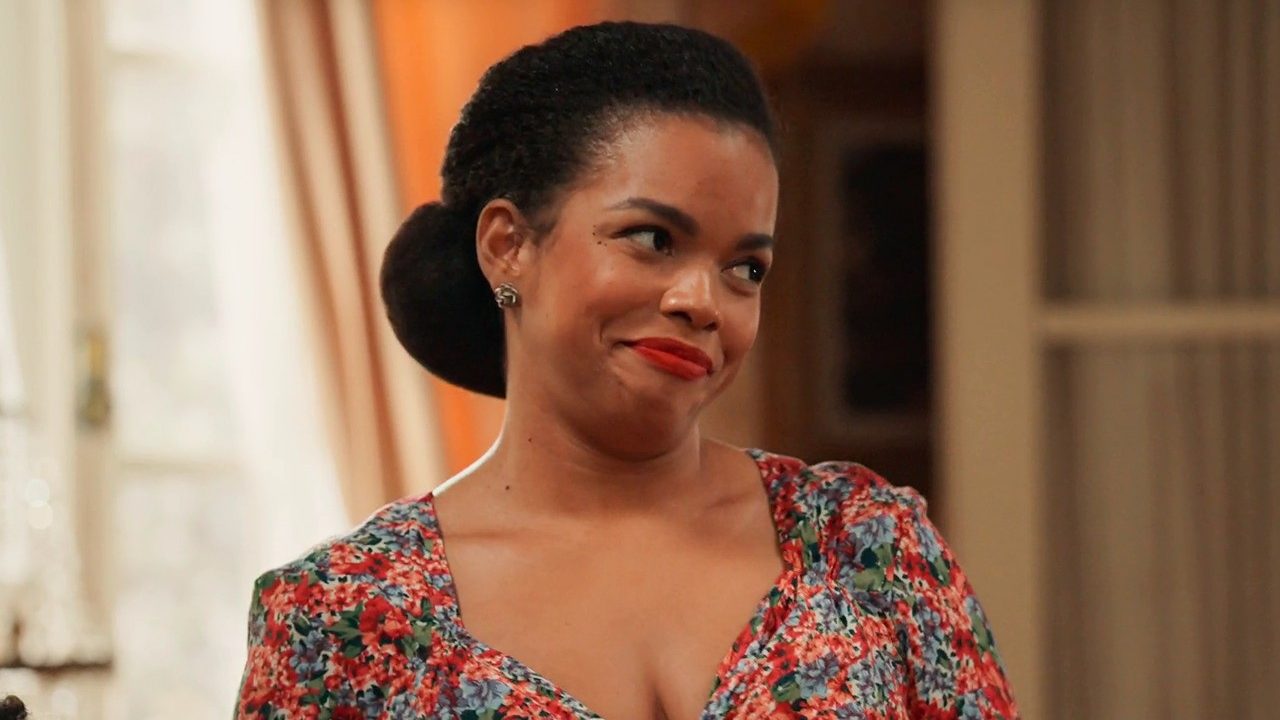 A imagem mostra uma mulher com expressão levemente divertida ou irônica, vestindo um vestido floral colorido e batom vermelho. Ela está em um ambiente interno bem iluminado, que parece ser uma sala decorada