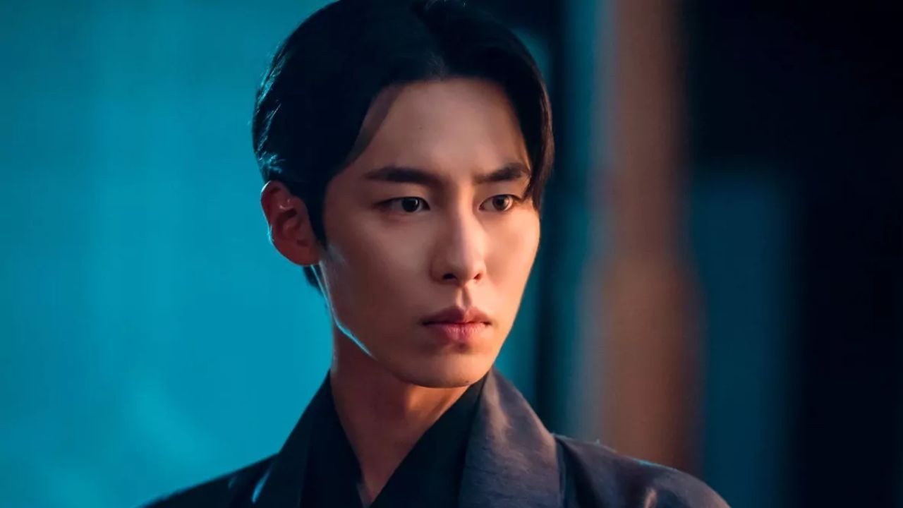 Homem de expressão séria olha para o lado em ambiente com iluminação azulada. Ele tem cabelo preto penteado para trás, usa camisa escura e está parcialmente iluminado por uma luz quente lateral, criando contraste entre sombras e tons frios ao fundo.